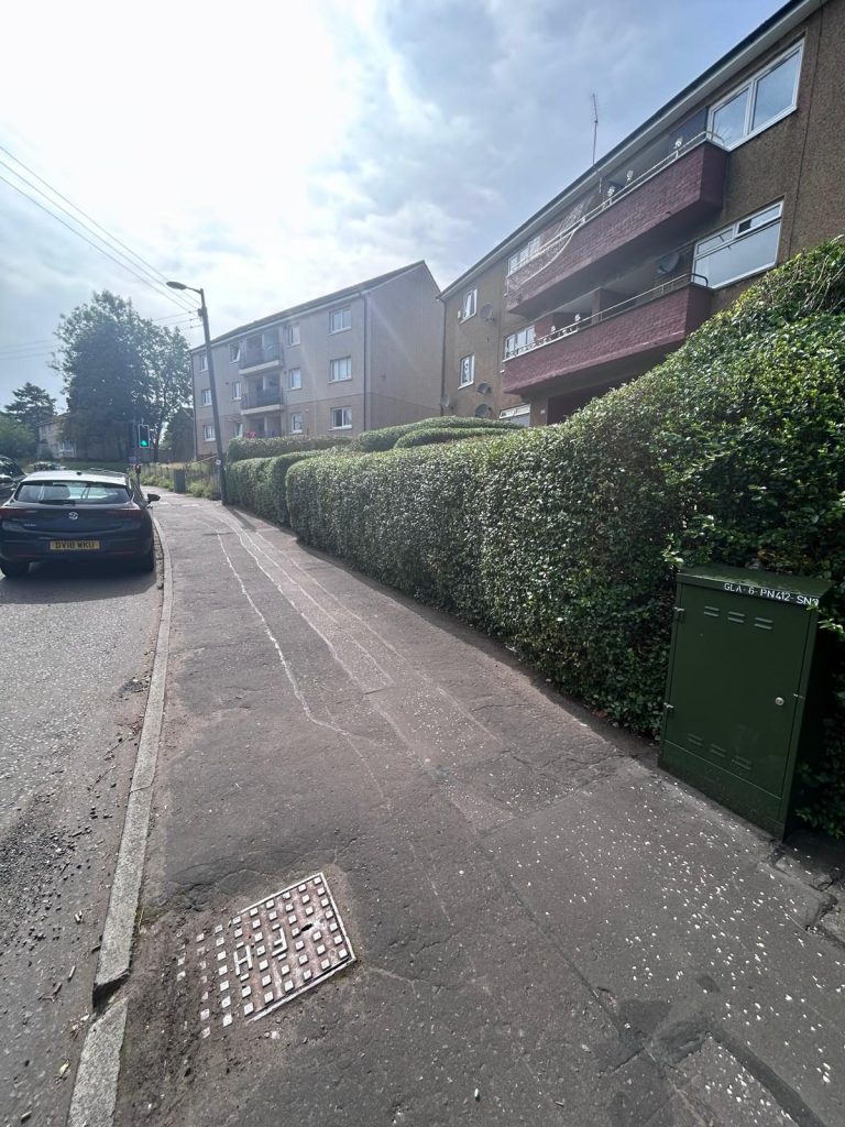 Residential street with green hedges, parked car, and nearby apartment buildings.